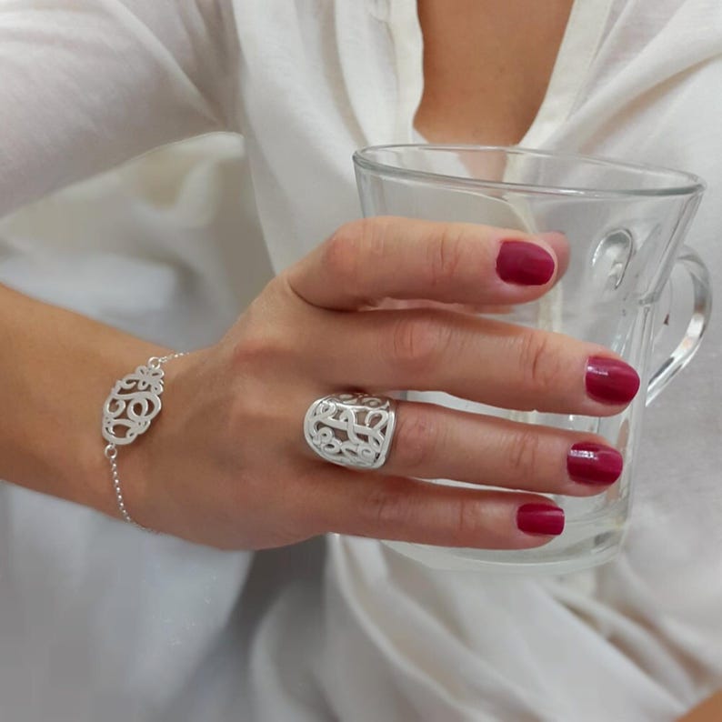 Op de afbeelding: Een zilveren armband- en ringenset. De armband heeft een opengewerkt ontwerp, verbonden door een fijne ketting. De bijpassende ring heeft een vergelijkbaar opengewerkt patroon. De nagels van de persoon zijn dieprood gelakt.