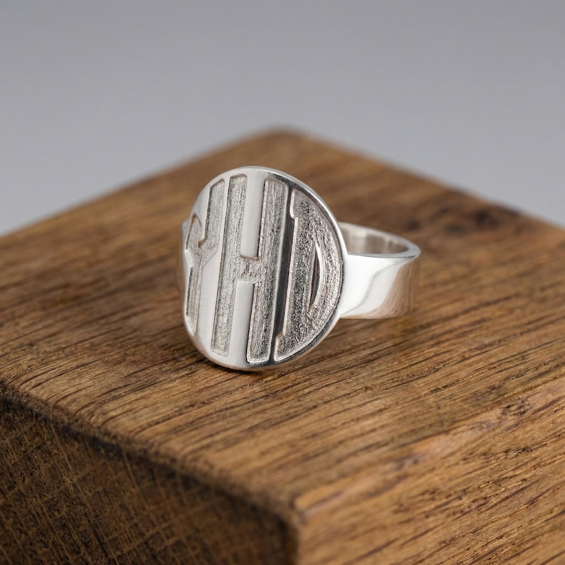 Op de afbeelding: Een zilveren zegelring met een groot, cirkelvormig oppervlak. Het oppervlak is gegraveerd met de letters "YHD" in een vetgedrukte, schreefloze letter. De ring ligt op een houten oppervlak.