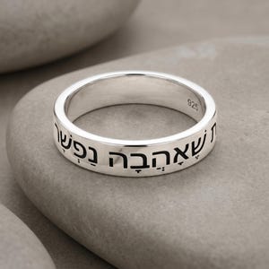 May include: A silver ring with Hebrew text engraved around the band. The ring is marked with "925" and sits on a gray stone surface. The ring's polished surface reflects light, highlighting the engraved letters.