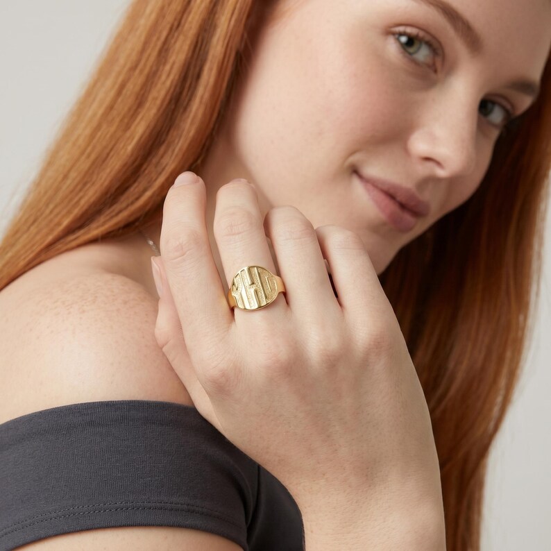 Op de afbeelding: Een goudkleurige zegelring met de initialen "HD" gegraveerd op de voorkant. De ring heeft een gladde band en een rechthoekige voorkant met afgeronde randen. De ring wordt om een vinger gedragen.
