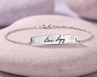 May include: A silver bracelet with a chain link design. The bracelet features a rectangular bar with the name "Eva Lopez" engraved in cursive script, along with a small heart symbol. The bracelet is displayed on a light purple stone.