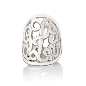 Op de afbeelding: Een zilverkleurige ring met een open ontwerp. De ring heeft een gestileerd monogram of initiaal, met de letters verweven in een ovale vorm. De ring staat tegen een witte achtergrond, met een lichte reflectie zichtbaar.