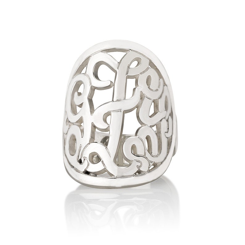 Op de afbeelding: Een zilverkleurige ring met een open ontwerp. De ring heeft een gestileerd monogram of initiaal, met de letters verweven in een ovale vorm. De ring staat tegen een witte achtergrond, met een lichte reflectie zichtbaar.