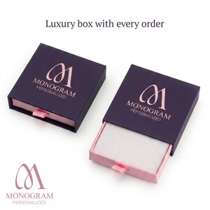 Op de afbeelding: Twee donkerpaarse geschenkdozen met roze interieurs en treklipjes. Elke doos heeft een ros&eacute;gouden monogramlogo en de woorden "MONOGRAM PERSONALIZED" op het deksel. De tekst "Luxury box with every order" is ook zichtbaar.