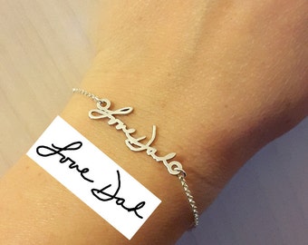 May include: A silver bracelet with the handwritten inscription "Love Dad" in cursive script. The bracelet is attached to a delicate chain. A white rectangle with the text "Love Dad" is in the lower left corner.