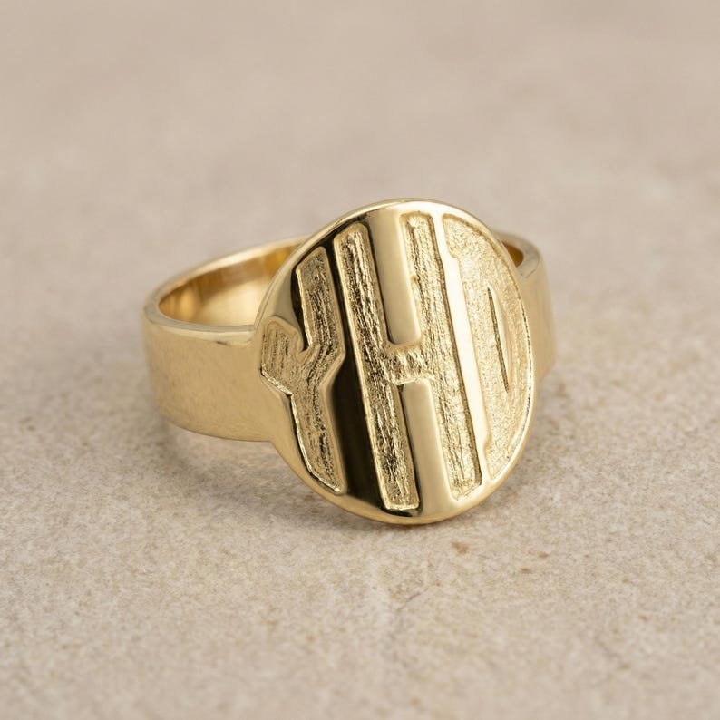 Op de afbeelding: Een goudkleurige zegelring met een grote, ronde voorkant. De voorkant heeft de letters "H O" in reli&euml;f in een vet, schreefloos lettertype. De ring heeft een dikke band en een gepolijste afwerking.