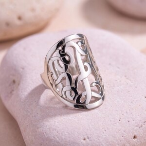 Op de afbeelding: Een zilverkleurige ring met een open ontwerp van verweven letters. De ring ligt op een lichtroze oppervlak. Het ontwerp van de ring is ingewikkeld en gedetailleerd, met een gepolijste afwerking.