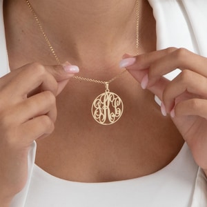 May include: A gold-colored pendant necklace featuring a circular design with an intricate monogram. The pendant hangs from a delicate chain. The necklace is being held up by a person.