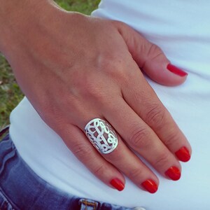 Op de afbeelding: Een zilverkleurige ring met een ingewikkeld, opengewerkt ontwerp met verweven letters. De ring is ovaal van vorm en wordt aan een vinger gedragen. De nagels van de persoon zijn rood gelakt.