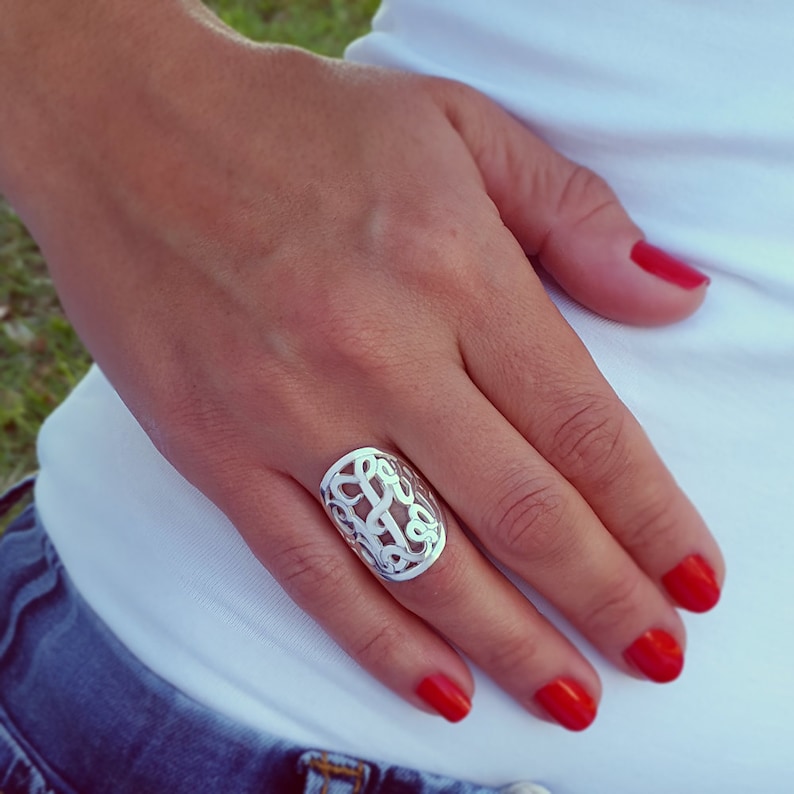 Op de afbeelding: Een zilverkleurige ring met een ingewikkeld, opengewerkt ontwerp met verweven letters. De ring is ovaal van vorm en wordt aan een vinger gedragen. De nagels van de persoon zijn rood gelakt.