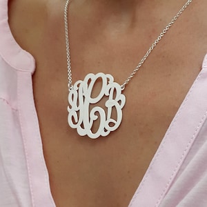 May include: A silver monogram necklace with the letters KPB in a decorative, intertwined design. The pendant hangs from a delicate chain. The necklace is displayed against a light pink background.