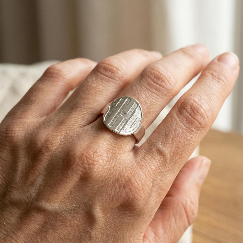 Op de afbeelding: Een zilveren zegelring met een cirkelvormige face met een gegraveerd ontwerp. De ring wordt aan een vinger gedragen en toont zijn gepolijste metalen afwerking. Het ontwerp lijkt een monogram of gestileerde initialen te zijn.