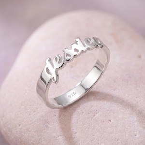 以下が含まれることがあります： 筆記体で「Jessie」と書かれたシルバーリング。リングは磨かれた金属製で、バンドの内側に「925」の数字が入っています。明るいピンク色の表面にリングが展示されています。