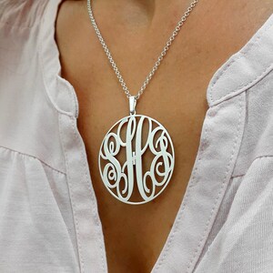K&ouml;nnte beinhalten: Eine silberne Halskette mit einem gro&szlig;en, runden Anh&auml;nger mit einem Monogramm-Design. Der Anh&auml;nger ist aus durchbrochenem Metall gefertigt und tr&auml;gt die Buchstaben "SFF" in einer kursivschriftlichen Schrift.