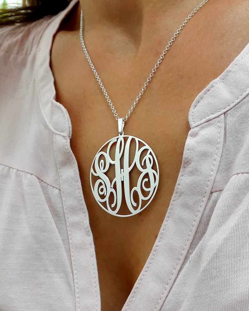 K&ouml;nnte beinhalten: Eine silberne Halskette mit einem gro&szlig;en, runden Anh&auml;nger mit einem Monogramm-Design. Der Anh&auml;nger ist aus durchbrochenem Metall gefertigt und tr&auml;gt die Buchstaben "SFF" in einer kursivschriftlichen Schrift.