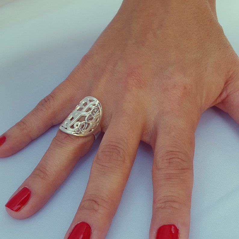 Op de afbeelding: Een zilverkleurige ring met een opengewerkt ontwerp wordt getoond op een hand met rode nagellak. De ring heeft een langwerpige, ovale vorm met ingewikkelde, wervelende patronen. De ring is een sieraad.