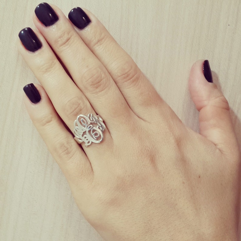Peut inclure: Une bague en argent avec un motif de monogramme. La bague est port&eacute;e sur un doigt avec un vernis &agrave; ongles noir.
