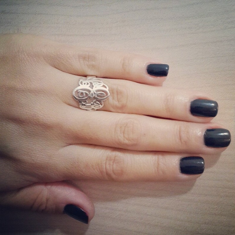 Peut inclure: Une bague en argent avec un motif de monogramme sur une main avec du vernis &agrave; ongles noir.