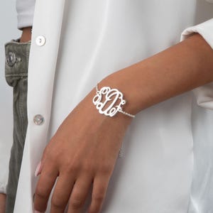 Könnte beinhalten: Ein silbernes Monogramm-Armband mit einem dreibuchstabigen Design. Das Armband ist durch eine filigrane Kette verbunden und wird am Handgelenk getragen. Der Hintergrund ist ein weißes Hemd.