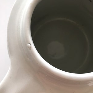 May include: White ceramic teapot with a smooth, glossy finish. The teapot has a round body and a spout on the side. The interior of the teapot is visible and is also white.