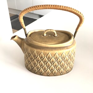 Può includere: Una teiera in ceramica marrone con un manico intrecciato e un motivo a foglie. La teiera ha un coperchio e un beccuccio.