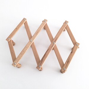 May include: A wooden magazine rack with a foldable design. The rack is made of light brown wood and has a natural finish. It is held together with silver metal rivets.