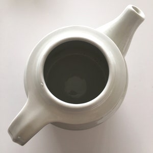 May include: White ceramic teapot with a rounded body and a spout. The teapot has a simple design and is empty.