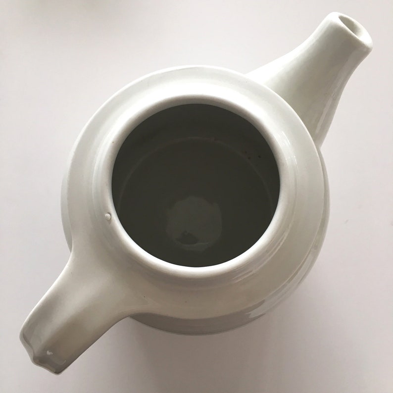May include: White ceramic teapot with a rounded body and a spout. The teapot has a simple design and is empty.