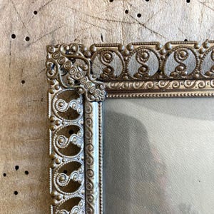 May include: A gold-toned ornate picture frame with a decorative, intricate design. The frame has a glass front and a smooth, light gray background.