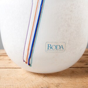 May include: White glass vase with a textured surface and a blue, purple, orange, and light blue stripe design. The vase has a label that reads "Boda Kosta Boda AB Sweden".