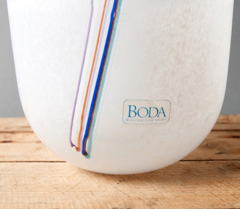 May include: White glass vase with a textured surface and a blue, purple, orange, and light blue stripe design. The vase has a label that reads "Boda Kosta Boda AB Sweden".