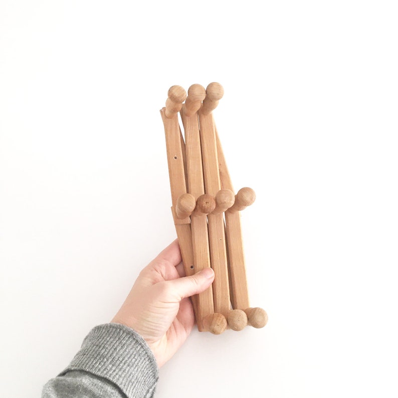 May include: A wooden coat rack with multiple hooks that folds up for storage. The rack is made of light brown wood and has a natural finish.