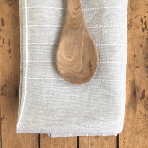 May include: A gray linen kitchen towel with white stitching and a wooden spoon on top.