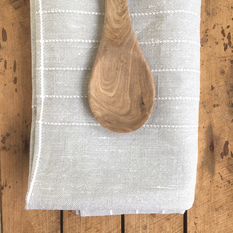 May include: A gray linen kitchen towel with white stitching and a wooden spoon on top.