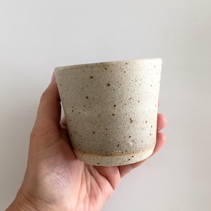 May include: A white ceramic cup with a speckled glaze. The cup has a rounded bottom and a slightly flared rim.