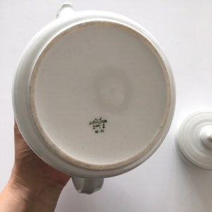 May include: White ceramic teapot with a lid. The bottom of the teapot has the text "ARABIA" in black lettering, "FINLAND" in smaller black lettering, and "10-71" in black lettering.
