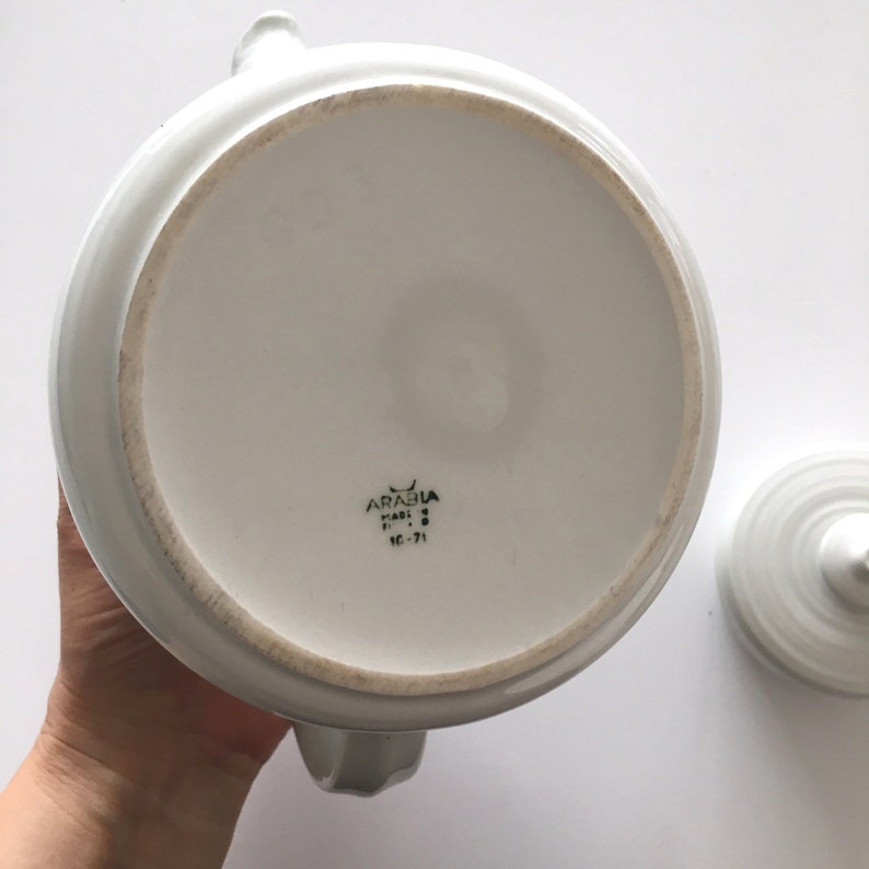May include: White ceramic teapot with a lid. The bottom of the teapot has the text "ARABIA" in black lettering, "FINLAND" in smaller black lettering, and "10-71" in black lettering.