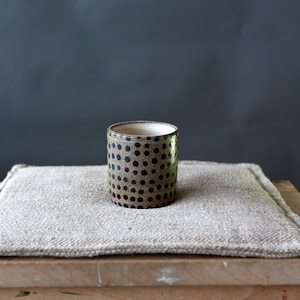 May include: A cylindrical ceramic cup with a speckled, tan background and evenly spaced dark blue polka dots. The cup sits on a beige, textured cloth, with a dark gray background. The cup is likely handmade.