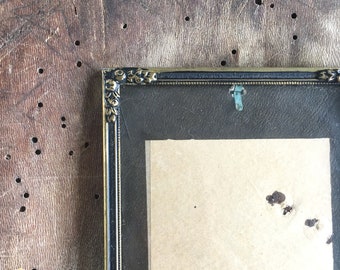 May include: A gold-toned picture frame with a black mount and a light brown paper insert. The frame has a decorative design around the edges.