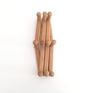 May include: A wooden coat rack with six hooks, folded up and ready to be hung on a wall. The rack is made of light brown wood and has a natural finish.
