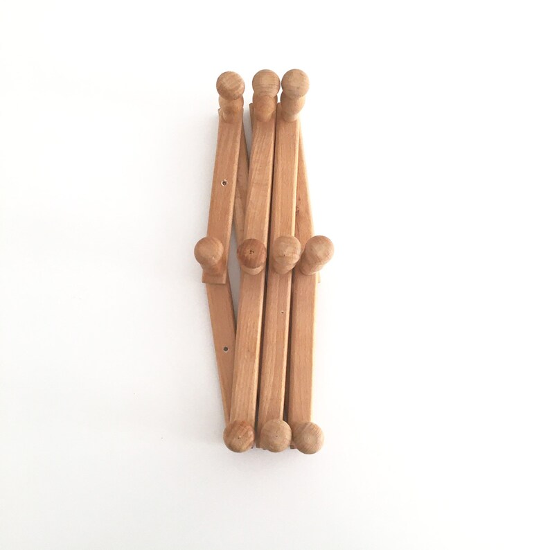 May include: A wooden coat rack with six hooks, folded up and ready to be hung on a wall. The rack is made of light brown wood and has a natural finish.
