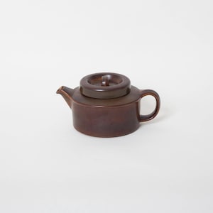 May include: A brown ceramic teapot with a lid. The teapot has a rounded body and a handle on the side.