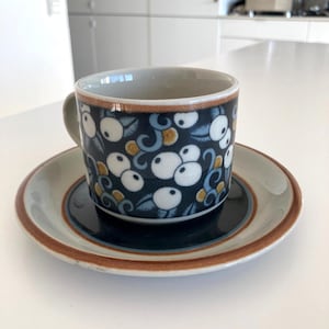Peut inclure: Un ensemble tasse et soucoupe à café en céramique. La tasse présente un fond bleu foncé avec un motif de motifs floraux blancs, bleus et dorés. La soucoupe a un design assorti avec un bord marron.