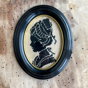 May include: A black and white embroidered silhouette of a person wearing a bonnet, framed in a black oval frame.
