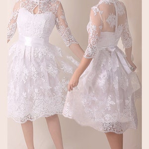 K&ouml;nnte beinhalten: Ein wei&szlig;es Spitzenkleid mit hohem Ausschnitt und einem figurbetonten Oberteil. Das Kleid hat einen vollen Rock und ein wei&szlig;es Satinband an der Taille. Das Kleid wird an einem Model mit wei&szlig;en Schuhen gezeigt.