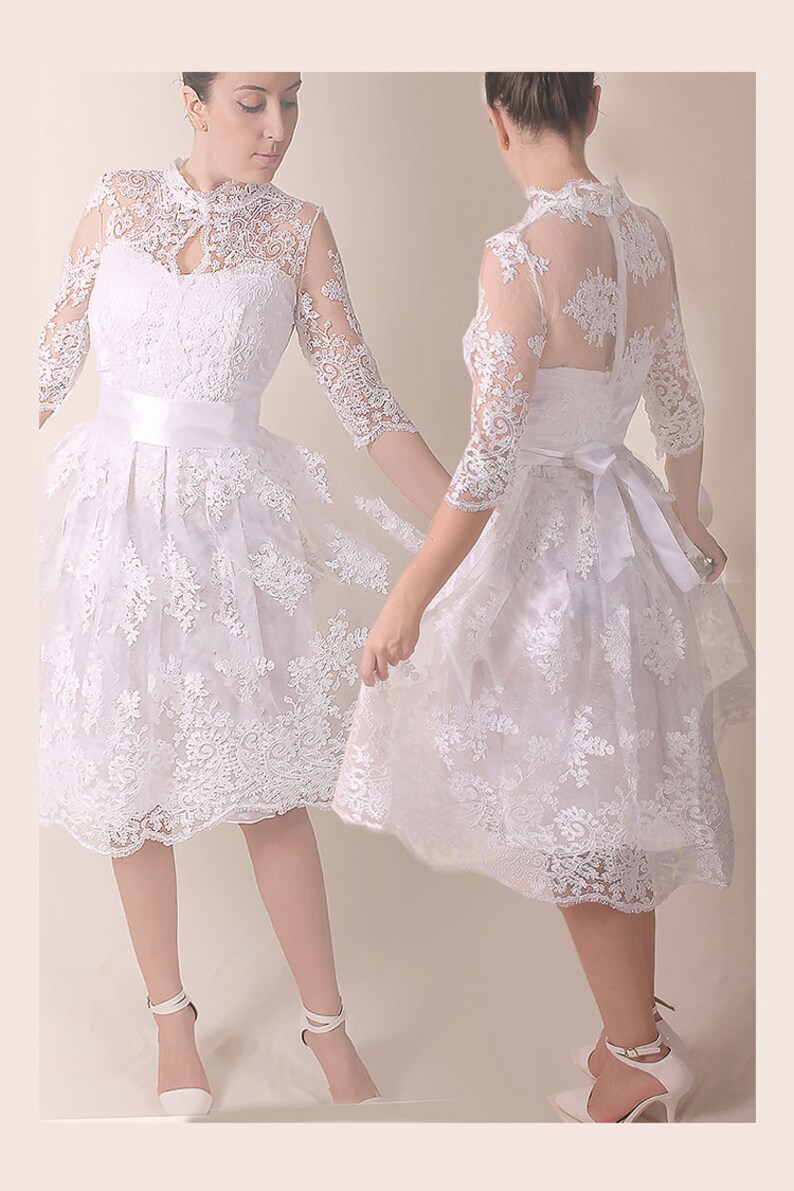 K&ouml;nnte beinhalten: Ein wei&szlig;es Spitzenkleid mit hohem Ausschnitt und einem figurbetonten Oberteil. Das Kleid hat einen vollen Rock und ein wei&szlig;es Satinband an der Taille. Das Kleid wird an einem Model mit wei&szlig;en Schuhen gezeigt.