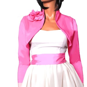 Puede incluir: Un bolero de satén rosa con una gran flor rosa en el hombro. El bolero tiene mangas largas y una longitud ligeramente recortada.
