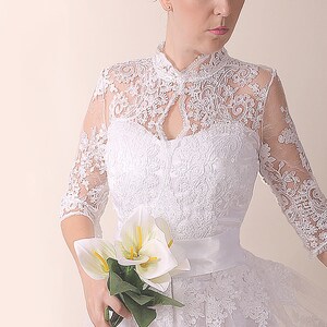 K&ouml;nnte beinhalten: Ein wei&szlig;es Spitzen-Hochzeitskleid mit hohem Ausschnitt und einem figurbetonten Oberteil. Das Kleid hat einen vollen Rock und eine Satinschleife. Die Braut h&auml;lt einen Strau&szlig; wei&szlig;er Calla-Lilien.