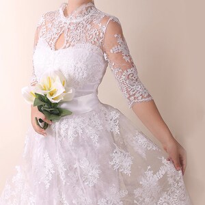 K&ouml;nnte beinhalten: Ein wei&szlig;es Spitzen-Hochzeitskleid mit hohem Ausschnitt und vollem Rock. Das Kleid hat eine Satinschleife in der Taille und ist mit zarter Blumenspitze verziert.