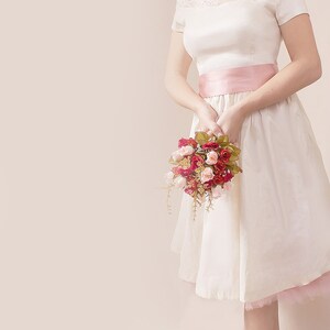 May include: A white dress with a lace bodice and a pink satin sash. The dress has a full skirt and is worn with a bouquet of pink and red roses.